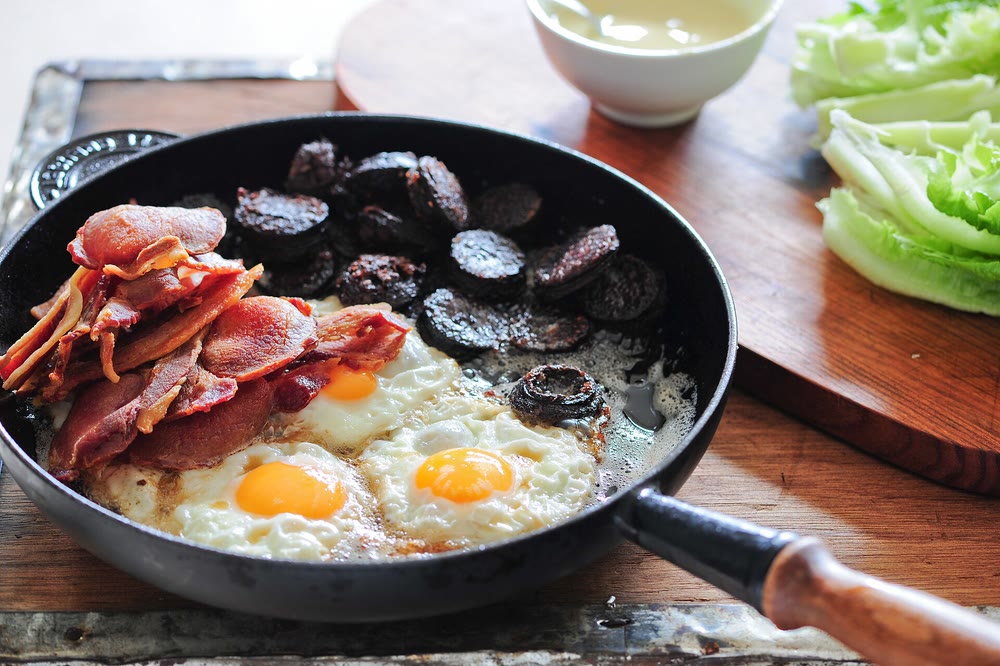 A black frying pan holding crisp rashers, slices of black pudding and three fried eggs, on a wooden board