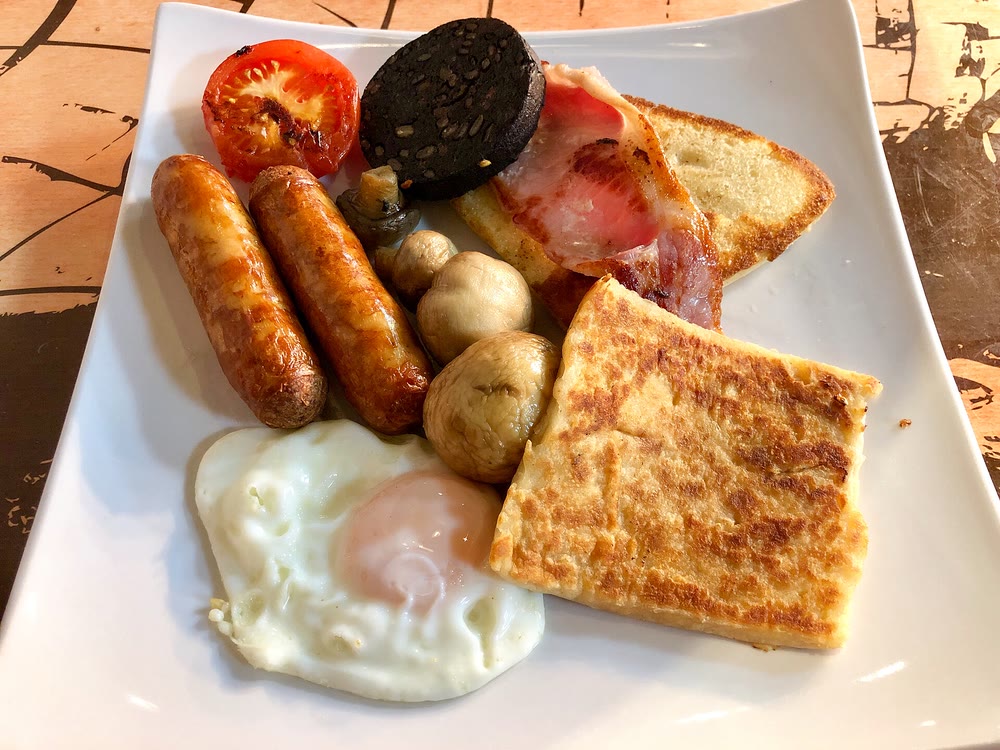 An Ulster fry on a square white plate: two sausages, a fried egg, mushrooms, grilled tomato, a round of black pudding, a rasher on fried soda bread and a square of golden potato bread