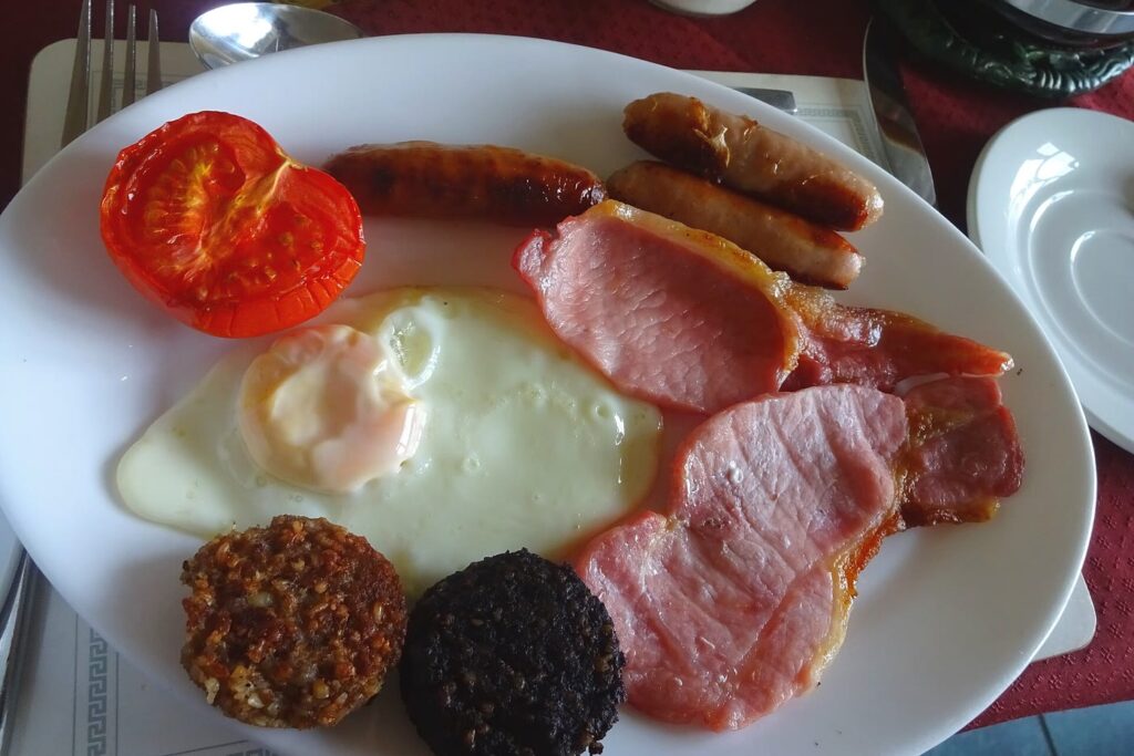 A full Irish breakfast on a white plate: half a grilled tomato, three pork sausages, back rashers, a fried egg and a round each of white and black pudding