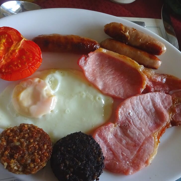 A full Irish breakfast on a white plate: half a grilled tomato, three pork sausages, back rashers, a fried egg and a round each of white and black pudding