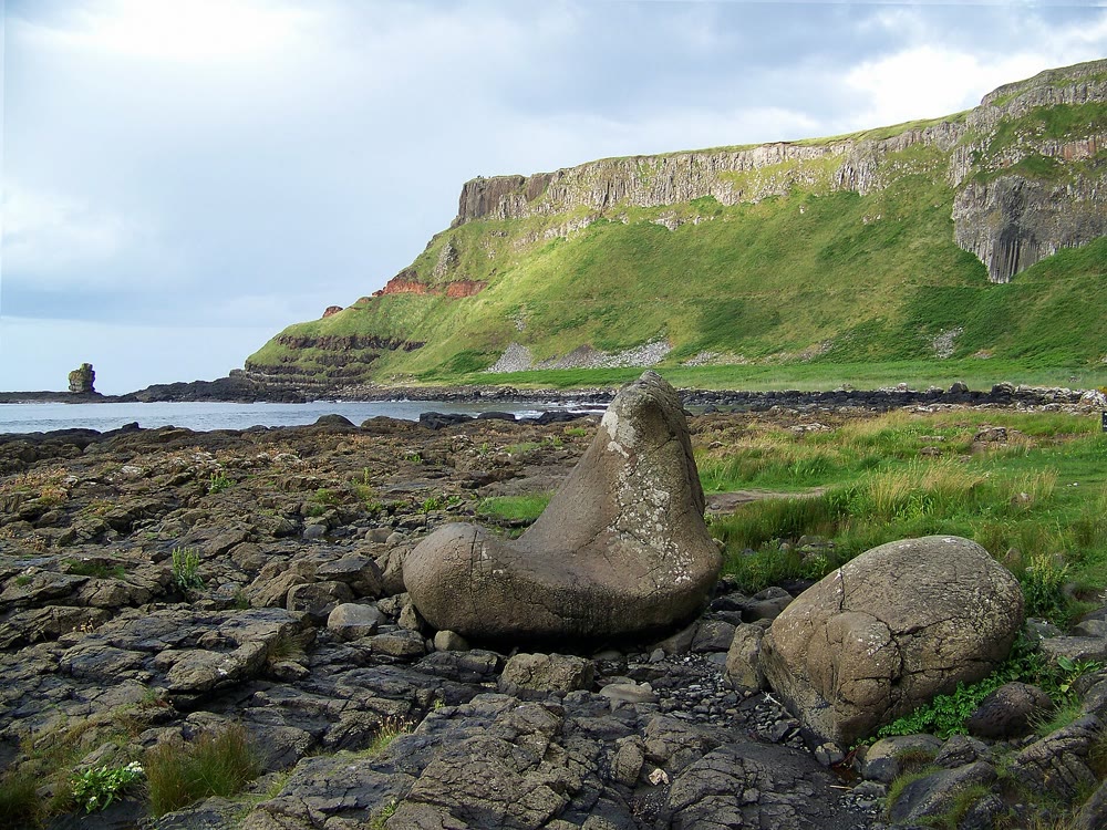 A large rounded boulder shaped like a boot sits on the rocky shore at Port Noffer, with green slopes and layered basalt cliffs rising behind it and the sea to the left
