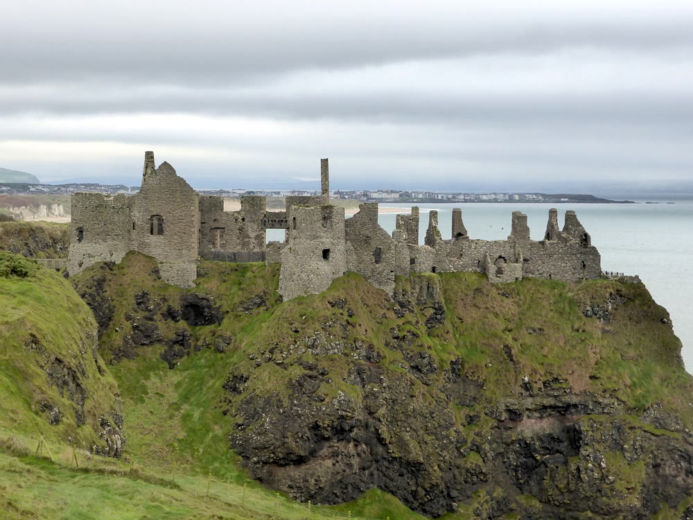 The ruined walls and gables of Dunluce Castle along the top of a grassy cliff above a dark rocky headland, with the sea and a distant coastal town under a grey sky
