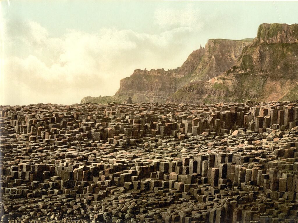 1890s colour photochrom of the Giant's Causeway: a broad pavement of stacked basalt columns in the foreground, with steep layered cliffs and headlands rising behind under a pale sky