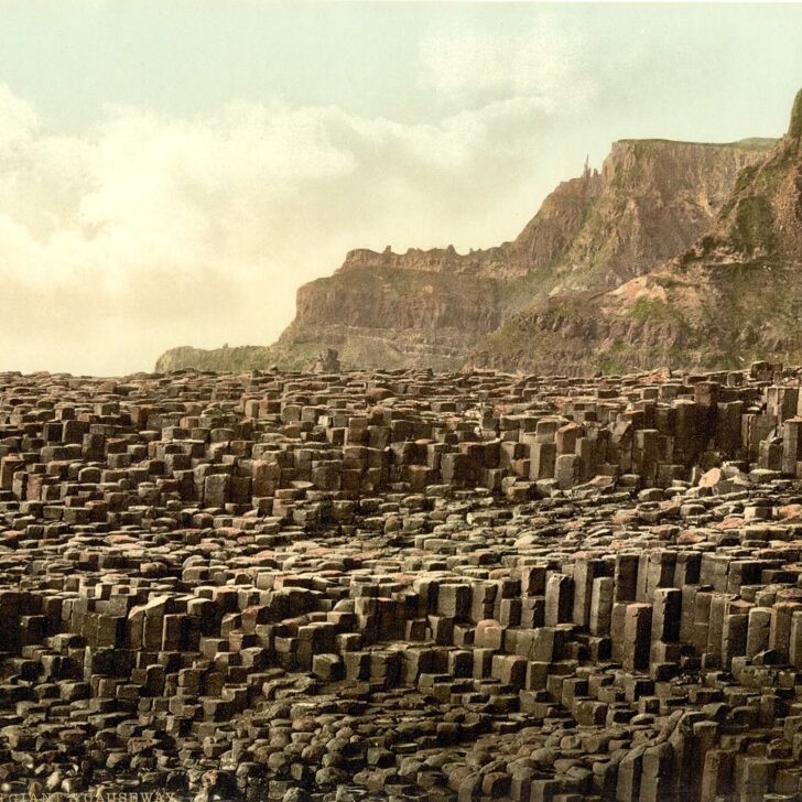 1890s colour photochrom of the Giant's Causeway: a broad pavement of stacked basalt columns in the foreground, with steep layered cliffs and headlands rising behind under a pale sky