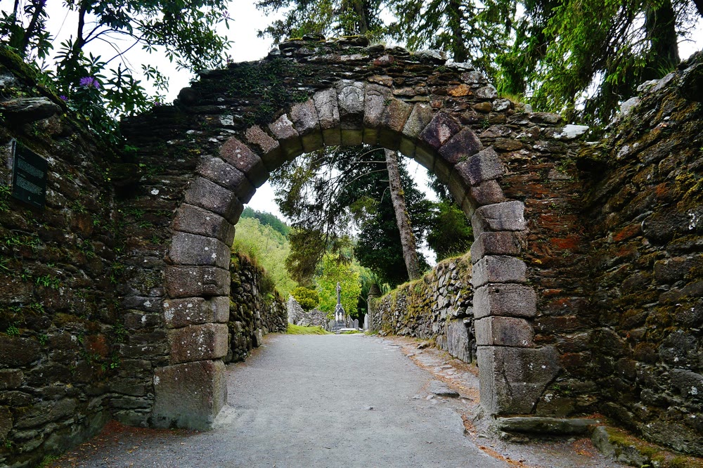 Glendalough gateway: a round arch of large granite blocks over a gravel path leading into the monastic city, with crosses and trees beyond