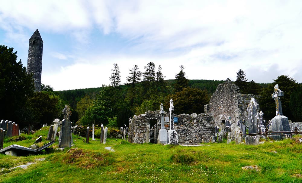 Glendalough round tower with its conical cap rising above the graveyard, with the roofless stone cathedral and Celtic crosses to the right