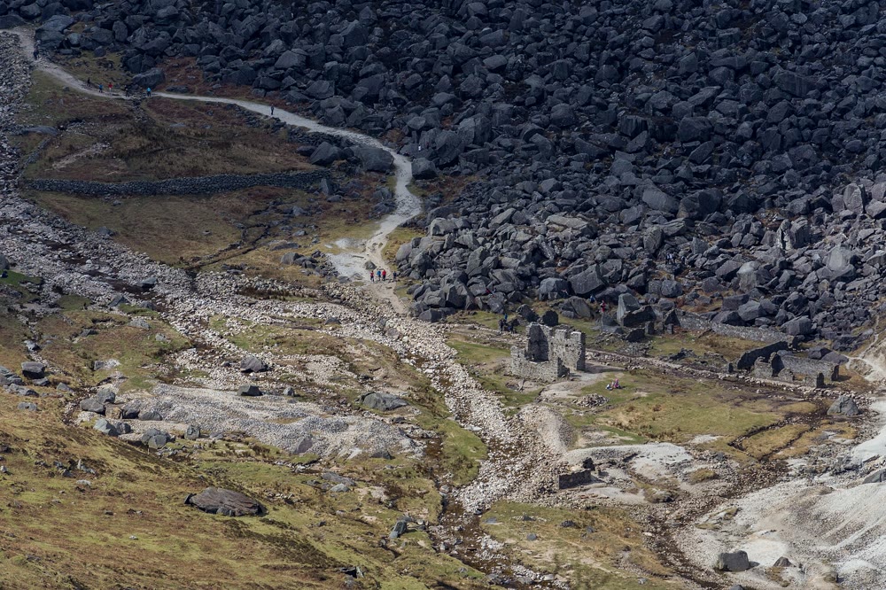 Ruined stone buildings of the Miners' Village at Glendalough among pale mine spoil, below a slope of dark boulders with a walking path winding uphill