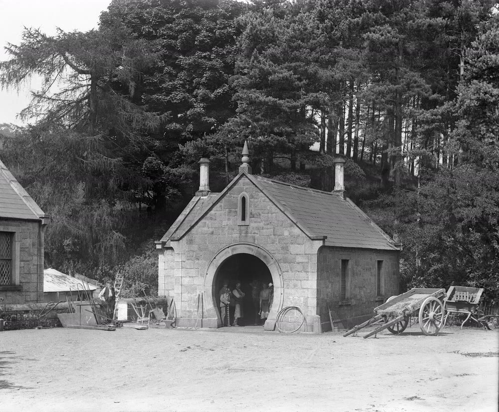 Black-and-white photograph of a small granite forge with a large horseshoe-shaped doorway, men standing in the opening, a cart and an iron wheel rim outside and tall trees behind