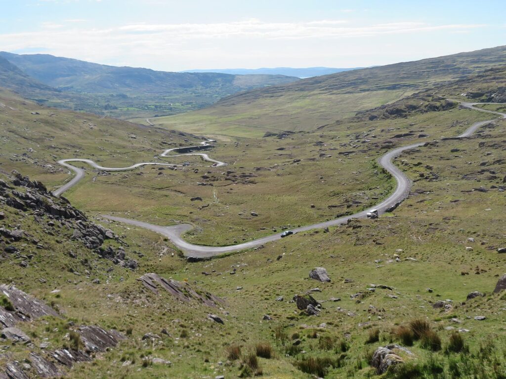 A narrow road zigzagging down a wide green mountain valley on the Healy Pass, with rocky slopes in front and hills and a glimpse of the sea in the distance