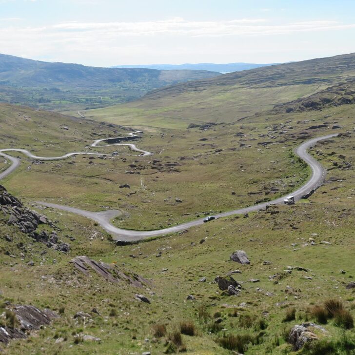 A narrow road zigzagging down a wide green mountain valley on the Healy Pass, with rocky slopes in front and hills and a glimpse of the sea in the distance