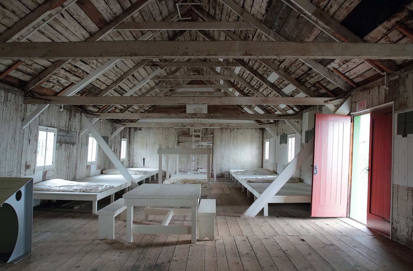 Inside the 1847 lazaretto at Grosse Île: rows of plain wooden bunks under a bare timber roof, with a red door open to the daylight