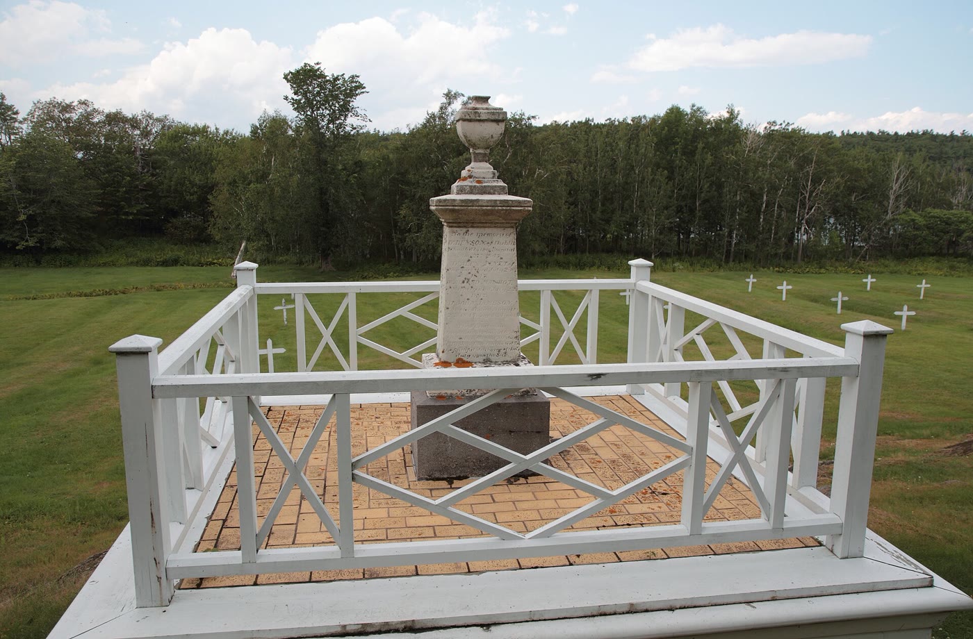 Weathered stone monument to the station's doctors, topped with an urn inside a white wooden railing, with small white crosses on the grass of the Irish cemetery behind it
