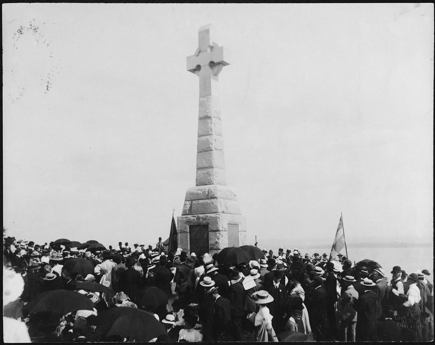 1909 photograph of a large crowd with umbrellas and straw hats gathered around the newly built granite Celtic cross on Grosse Île, with flags flying
