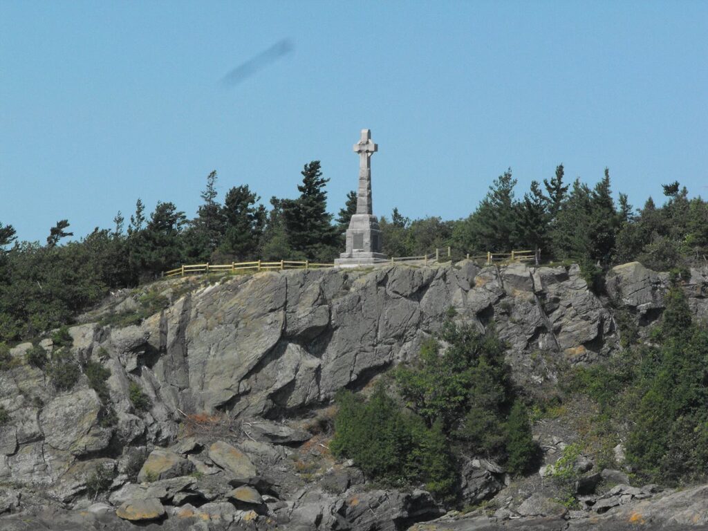The 1909 Celtic cross memorial at Grosse Île, Quebec, standing on a rocky cliff top among evergreen trees under a clear blue sky