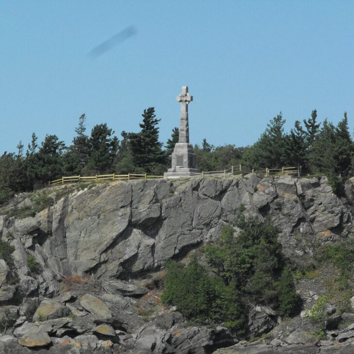 The 1909 Celtic cross memorial at Grosse Île, Quebec, standing on a rocky cliff top among evergreen trees under a clear blue sky