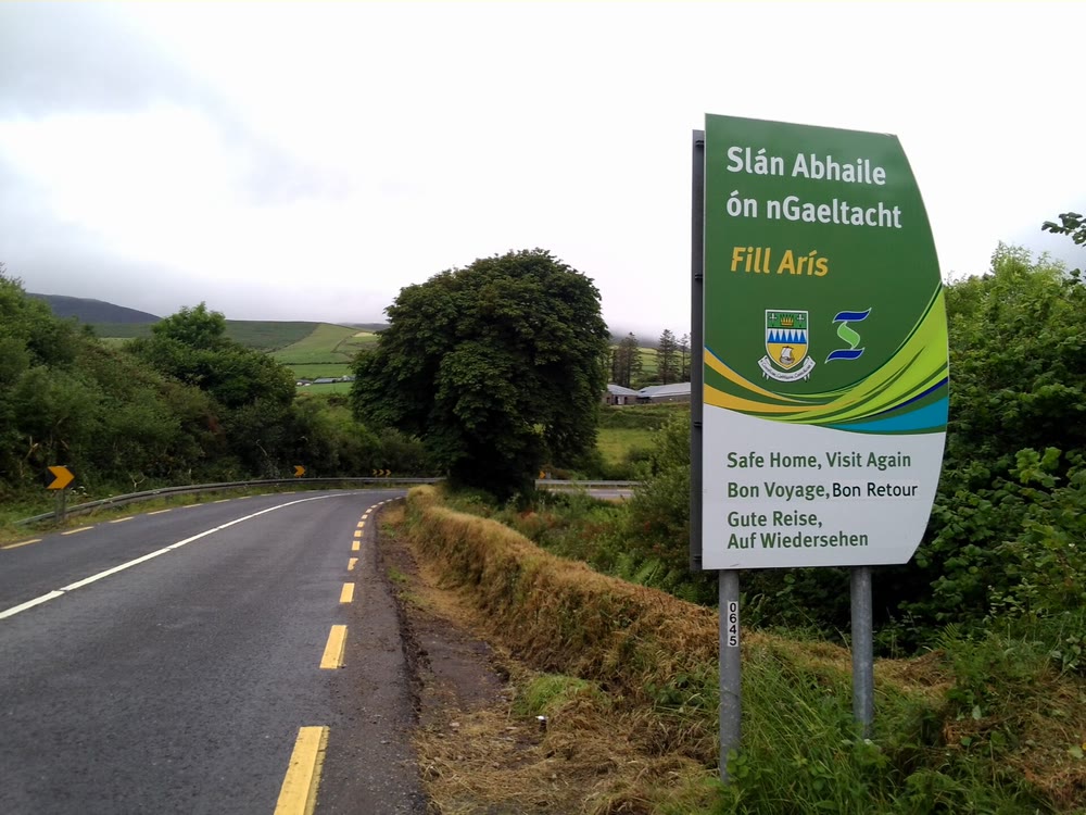 Green and white roadside sign reading Sl&aacute;n Abhaile &oacute;n nGaeltacht, Fill Ar&iacute;s, with Safe Home, Visit Again in English below, beside a country road with hedges and green fields