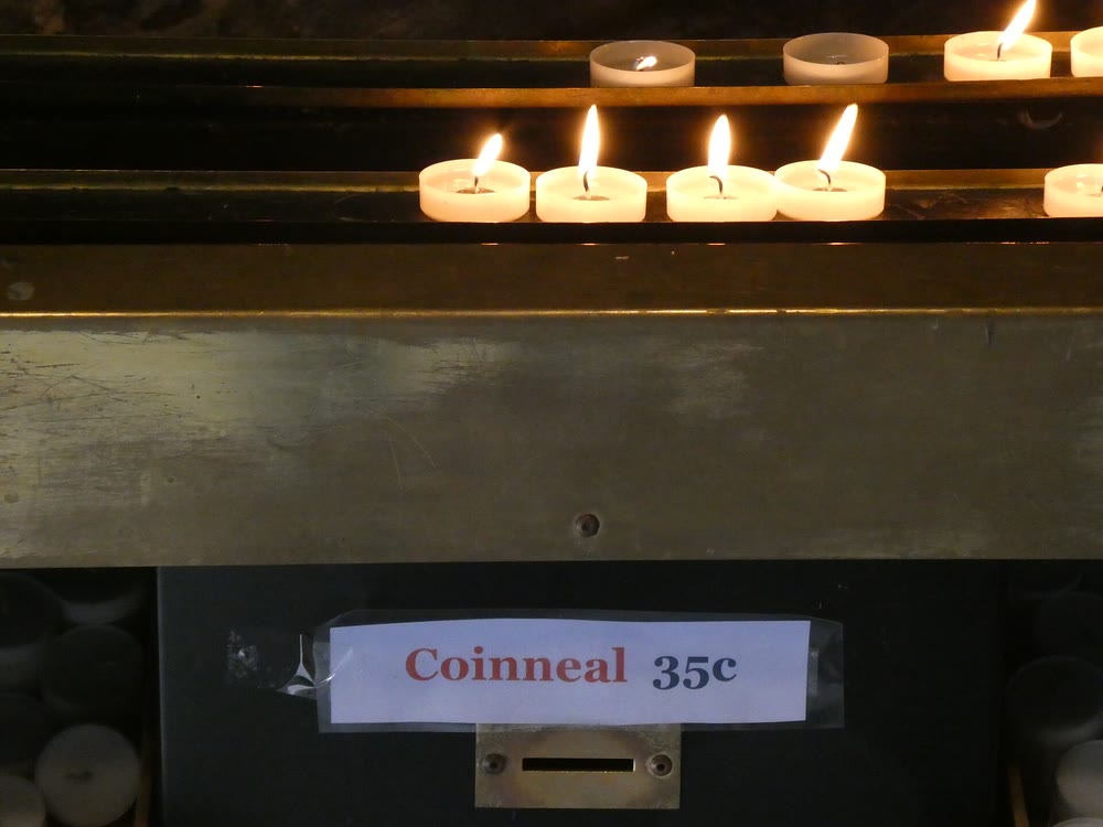 Lit tea-light candles on a brass stand above a small printed sign reading Coinneal 35c