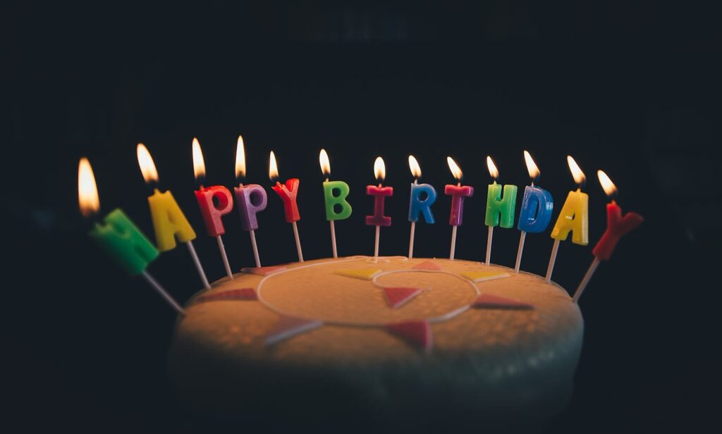 Birthday cake on a dark background with lit, brightly coloured letter candles spelling HAPPY BIRTHDAY