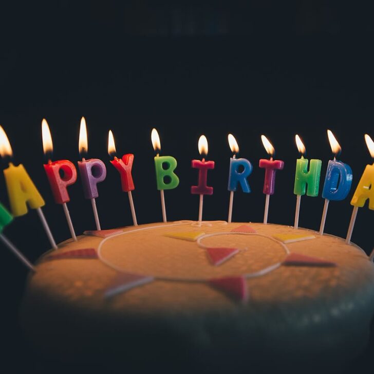 Birthday cake on a dark background with lit, brightly coloured letter candles spelling HAPPY BIRTHDAY
