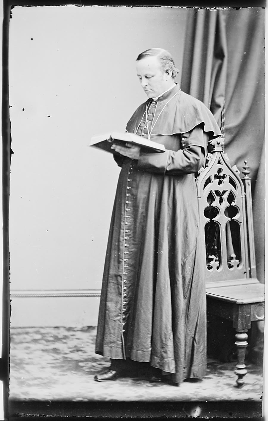Full-length black and white photograph of Archbishop John Hughes of New York in clerical robes, reading a book