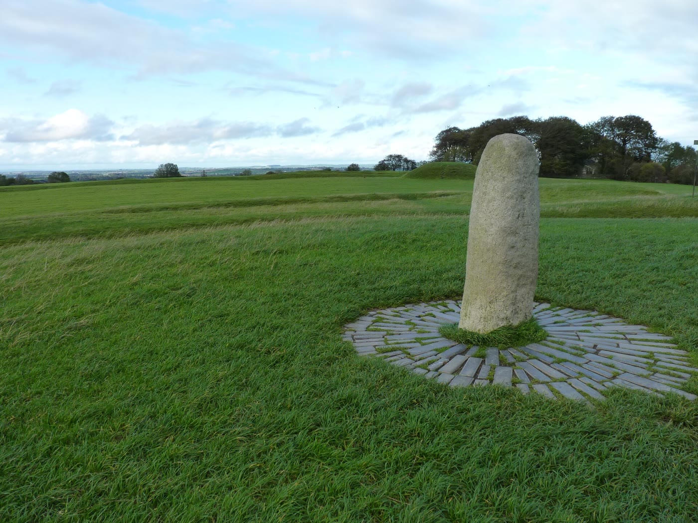The Lia F&aacute;il on the Hill of Tara: a rounded grey standing stone in a ring of slate paving on a grassy mound, with earthwork banks and trees behind