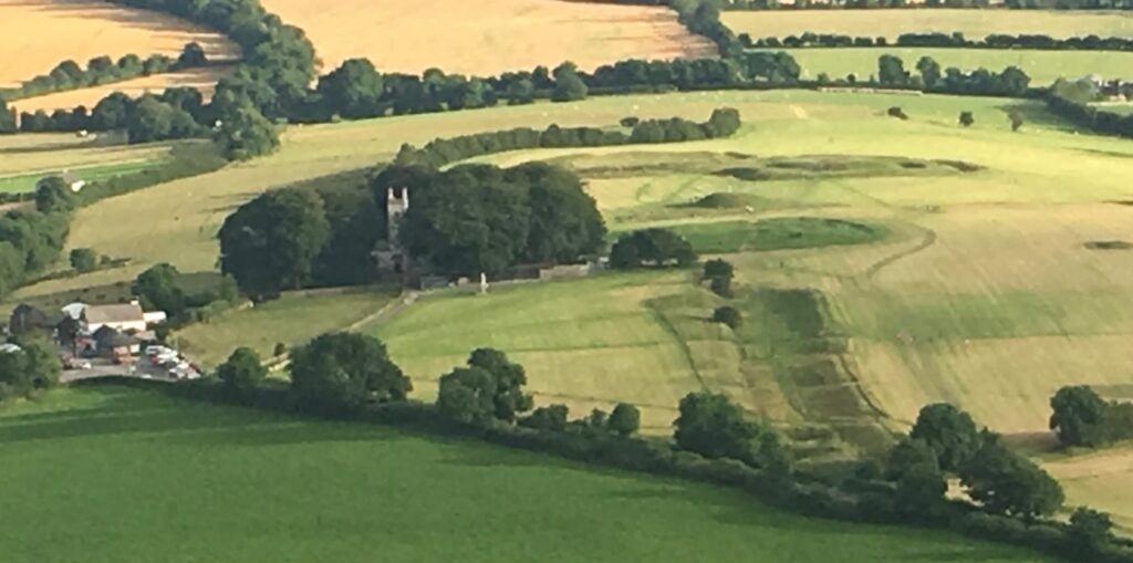 The Hill of Tara from the air: grassy earthwork rings and banks on the ridge beside St Patrick's Church and its tower among trees, with green fields beyond