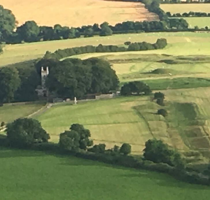 The Hill of Tara from the air: grassy earthwork rings and banks on the ridge beside St Patrick's Church and its tower among trees, with green fields beyond