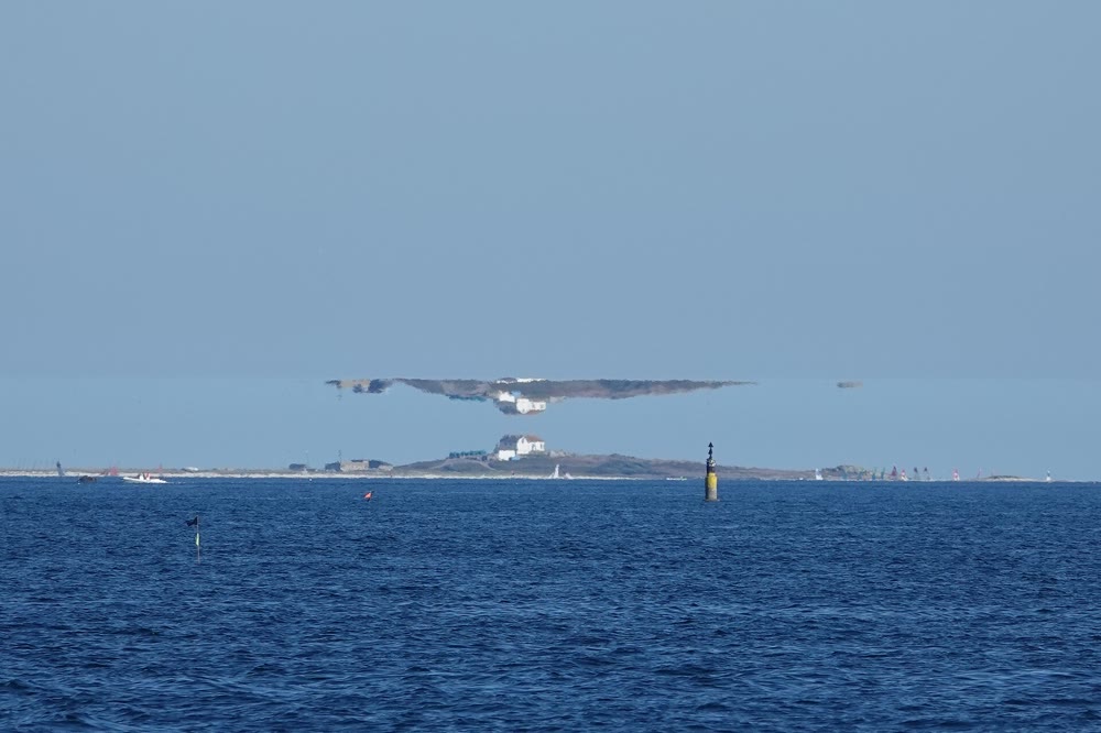 A low island with a white house on the horizon of a calm blue sea, with a stretched, upside-down mirage of the island hovering in the sky just above it