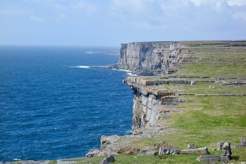 Sheer grey limestone cliffs on Inis Mór dropping into a deep blue Atlantic, with green clifftop fields and old stone walls under a pale sky