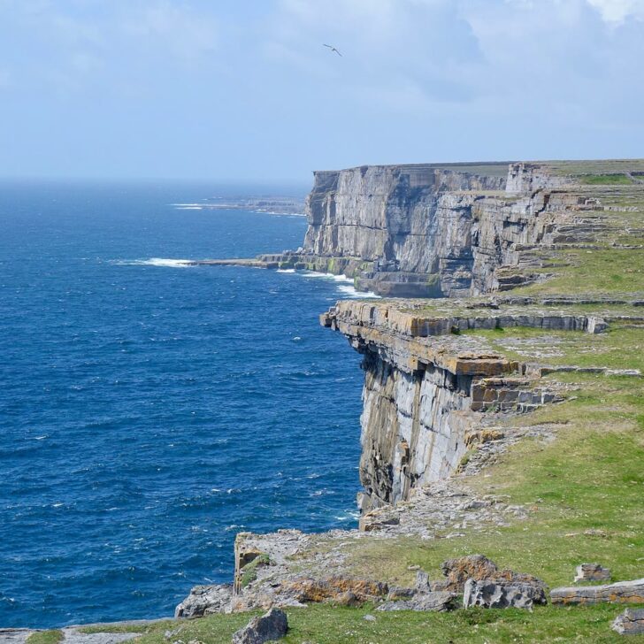 Sheer grey limestone cliffs on Inis Mór dropping into a deep blue Atlantic, with green clifftop fields and old stone walls under a pale sky