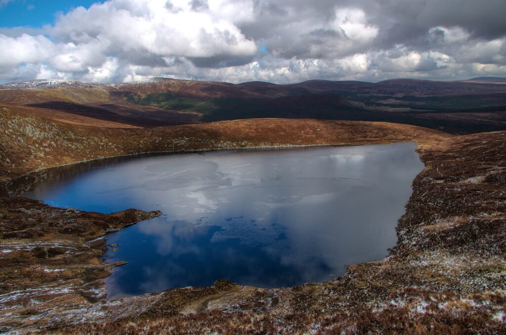 Lough Ouler, a heart-shaped mountain lake reflecting white clouds, set among brown heather slopes lightly dusted with snow in the Wicklow Mountains