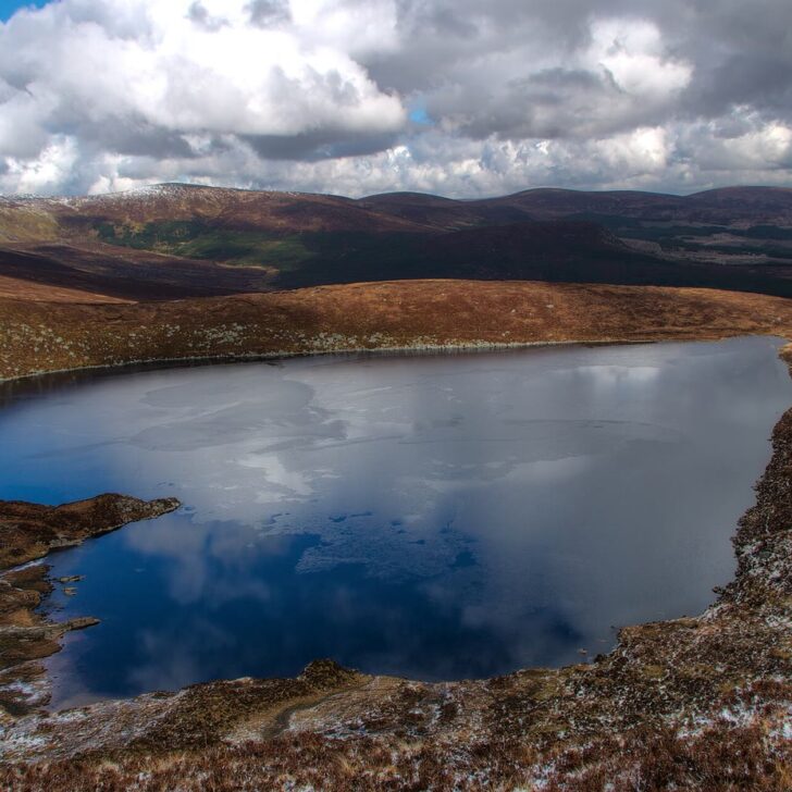 Lough Ouler, a heart-shaped mountain lake reflecting white clouds, set among brown heather slopes lightly dusted with snow in the Wicklow Mountains