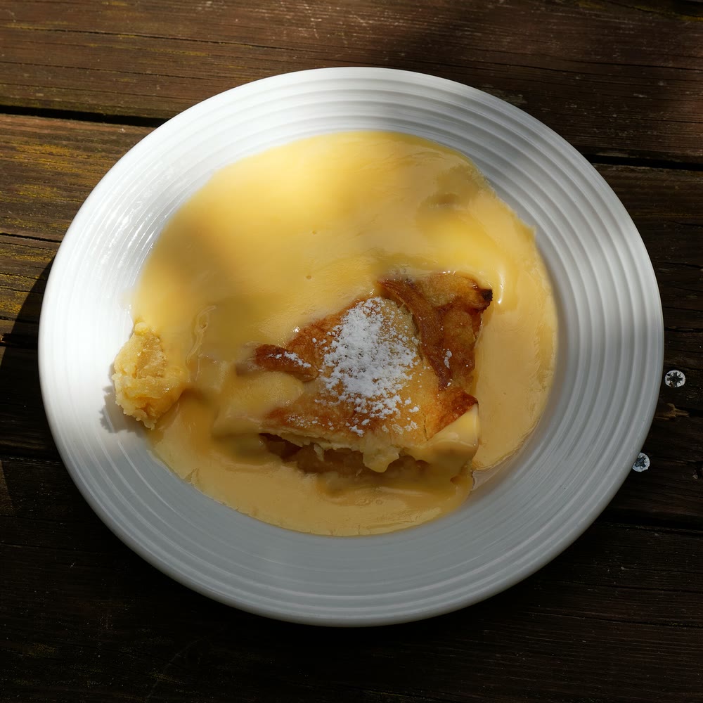 A piece of golden apple tart dusted with icing sugar, sitting in a pool of yellow custard in a white bowl on a wooden table