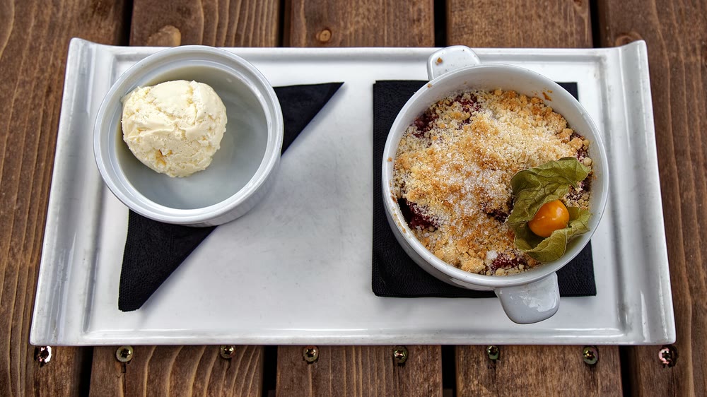 A white dish of blackberry and apple crumble with a sugary golden topping and a physalis garnish, beside a small bowl of vanilla ice cream on a white tray