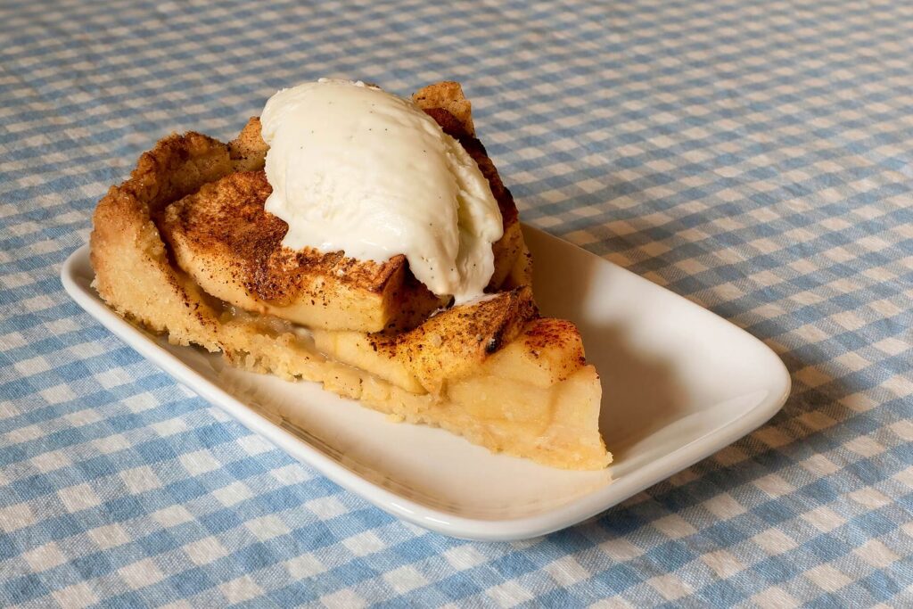 A slice of apple bake topped with cinnamon-dusted baked apple slices and a scoop of vanilla ice cream, on a white plate on a blue gingham tablecloth