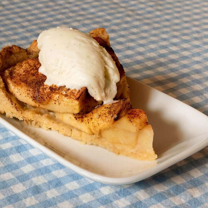 A slice of apple bake topped with cinnamon-dusted baked apple slices and a scoop of vanilla ice cream, on a white plate on a blue gingham tablecloth