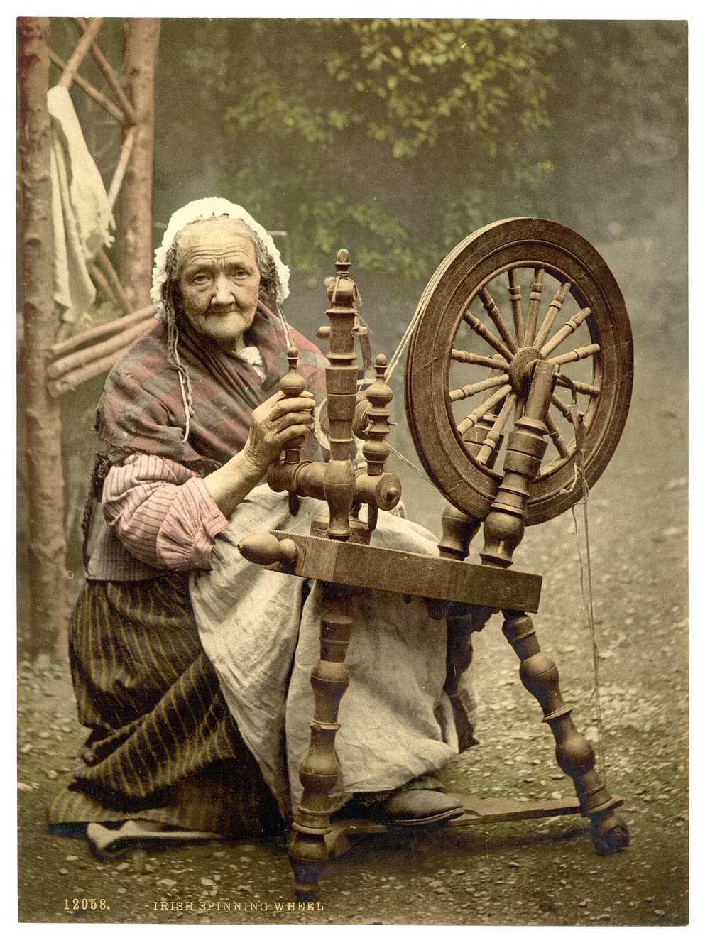 Hand-coloured 1890s photochrom of an elderly woman in a white cap and shawl sitting at a wooden spinning wheel in Co. Galway