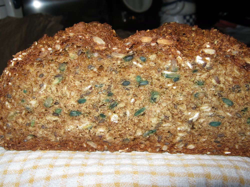 Close-up of the cut face of a loaf of brown soda bread, its crumb flecked with oats, linseed and green-tinged seeds, resting on a yellow and white checked tea towel