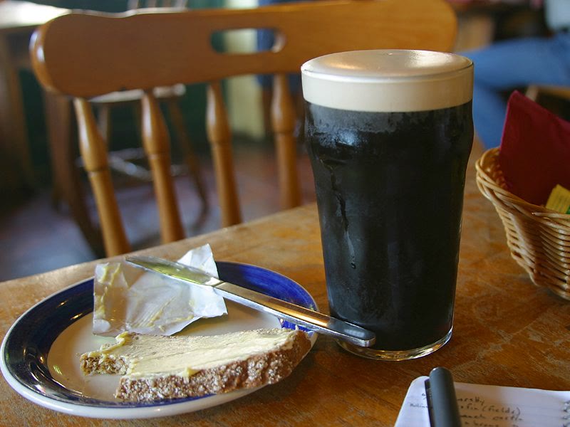 A pint of stout with a creamy head on a wooden pub table, beside a blue-rimmed plate holding a slice of buttered brown bread and a butter knife