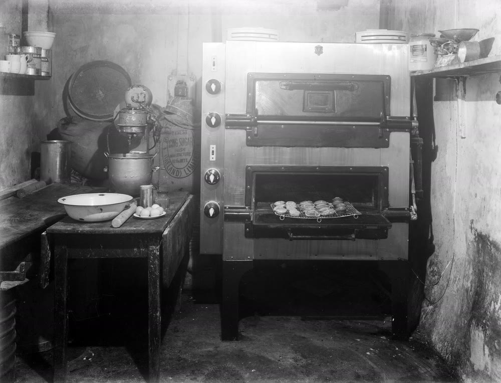 A black-and-white photograph of a 1940s baking room: a large two-door electric oven with a tray of buns at its open lower door, beside a table with an enamel basin, rolling pin, eggs and a mixer, and sacks stacked against the wall