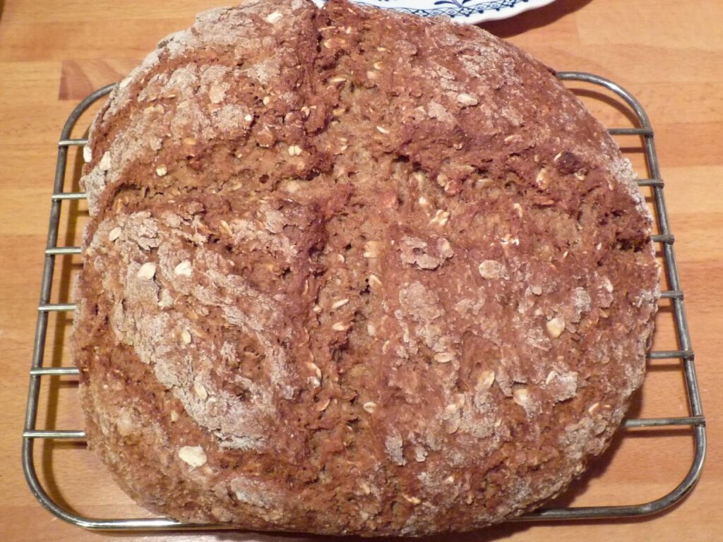 A round loaf of brown soda bread studded with oats and dusted with flour, with a shallow cross in the top, cooling on a wire rack on a wooden worktop
