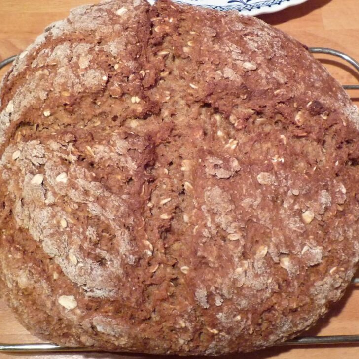 A round loaf of brown soda bread studded with oats and dusted with flour, with a shallow cross in the top, cooling on a wire rack on a wooden worktop