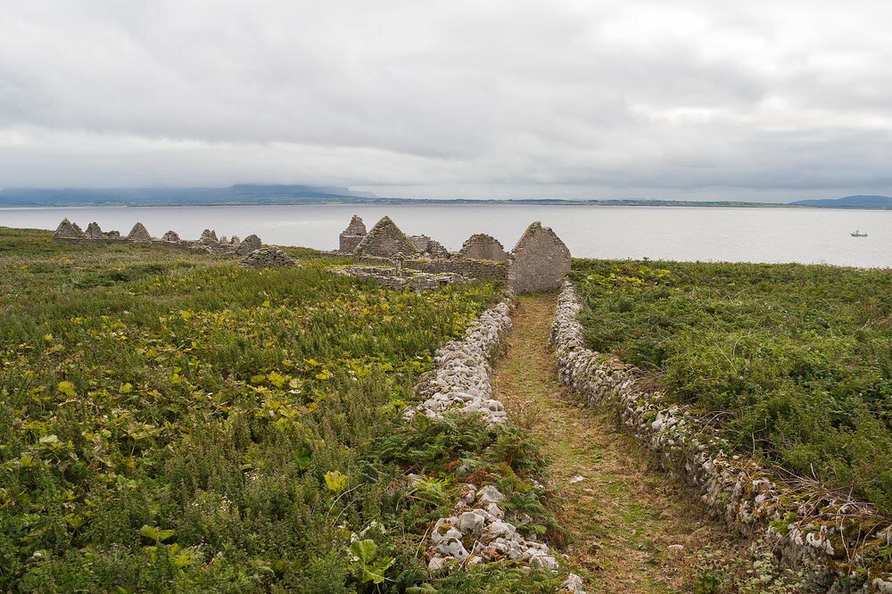 A grassy path between low stone walls on Inishmurray leading towards roofless stone gables, with grey sea and distant mountains under a cloudy sky