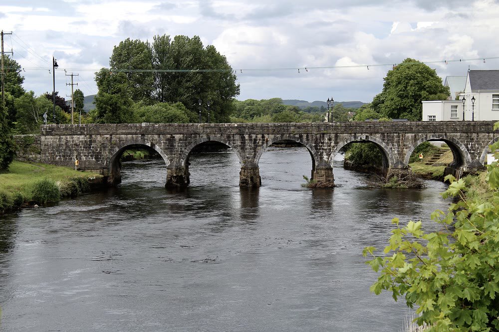 A long stone bridge with several arches crossing the dark River Moy at Foxford, with trees and a white house on the far bank