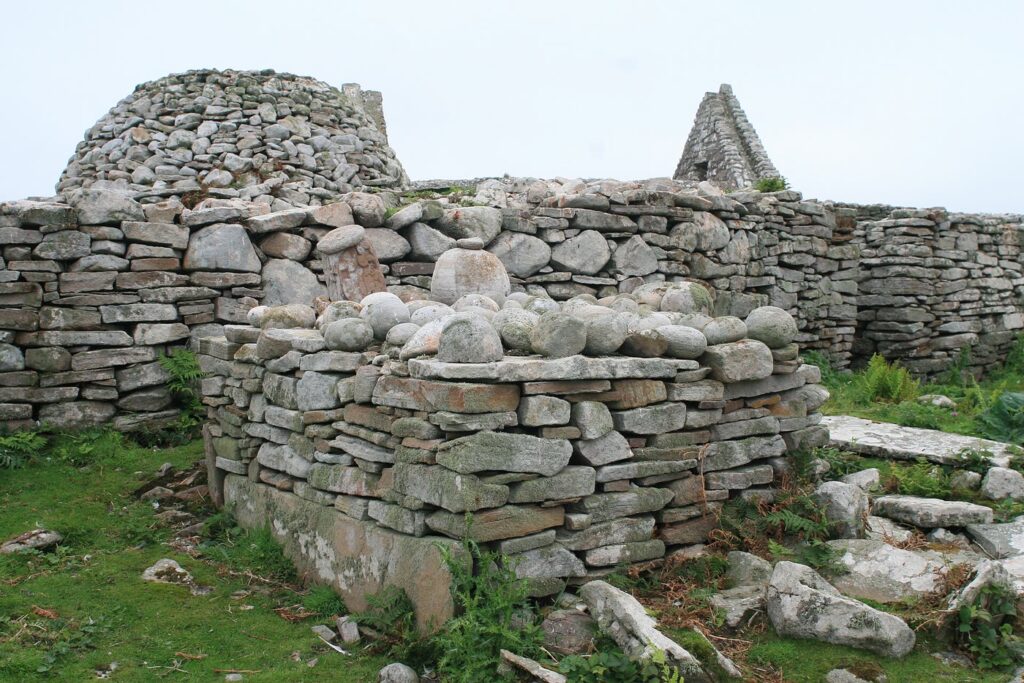 The Clocha Breaca on Inishmurray: rounded cursing stones heaped on a low drystone altar, with a stone beehive cell and a ruined church gable behind