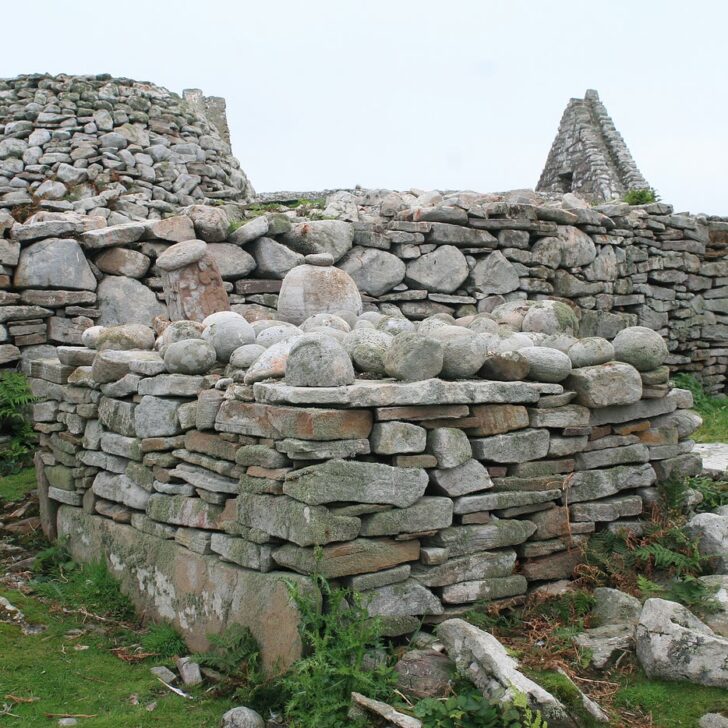 The Clocha Breaca on Inishmurray: rounded cursing stones heaped on a low drystone altar, with a stone beehive cell and a ruined church gable behind