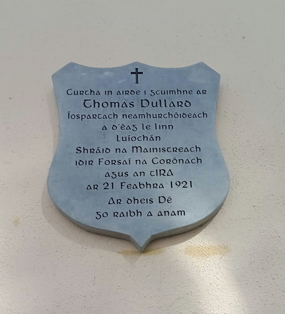 Grey shield-shaped stone plaque on a cream wall, with a carved cross and Irish text in Gaelic lettering ending with the words Ar dheis D&eacute; go raibh a anam