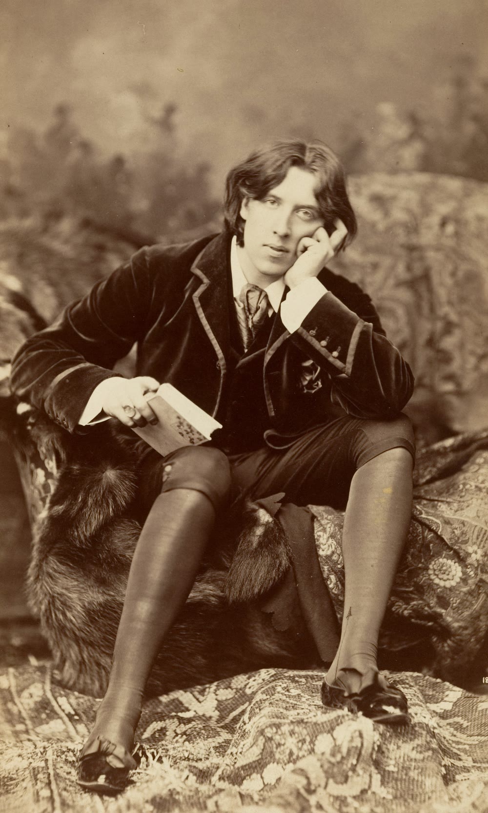 Oscar Wilde in 1882, seated on a draped couch with his chin resting on one hand and a book in the other, wearing a velvet jacket, knee breeches and stockings