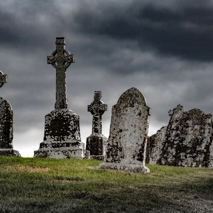Weathered Celtic cross headstones and old gravestones on a grassy rise at Clonmacnoise, Co. Offaly, under a dark, cloudy sky