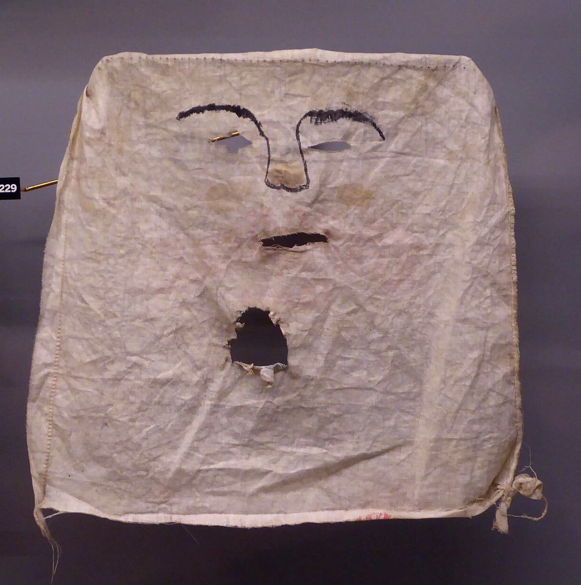Old Irish Halloween mask made from a square of creased white flour-bag cloth, with cut-out eyes and mouth and painted black eyebrows and nose, displayed in a museum case
